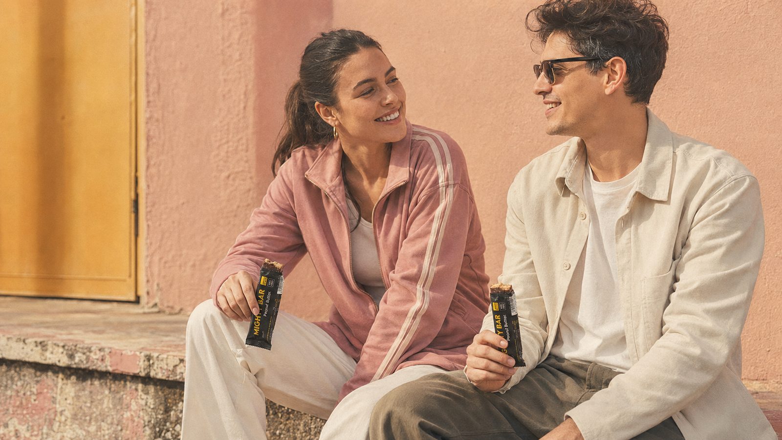 A man and a woman sitting outdoors, smiling, each holding a Mighty Bar peanut butter protein bar